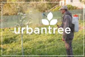 A person wearing a backpack sprayer and a cap is spraying a small, blossoming tree in a grassy area. The "urbantree" logo is overlaid in white on the image.