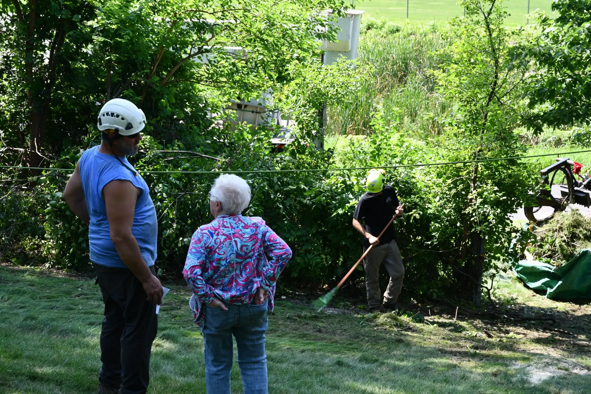 Urban Tree crew lead in a white climbing helmet standing with a homeowner while a crew member rakes up brush behind them