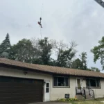 Climber suspended from the crane above a Blaine roof while preparing for crane tree removal